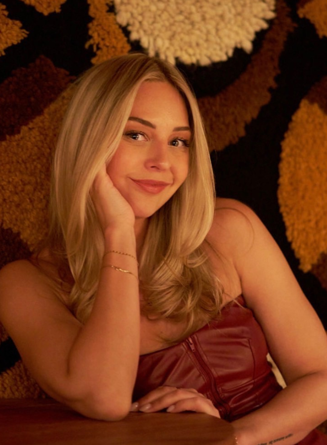 Young woman with long blonde hair resting her head on her hand, smiling softly, wearing a sleeveless burgundy top with a zipper, sitting at a wooden table with a textured, multicolored background.